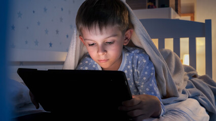 Portrait of little boy lying under blanket and using tablet computer. Children education, development, kids using gadgets secrecy, privacy