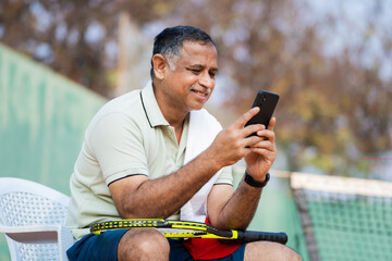 Happy senior man at tennis court busy using mobile phone during break while playing game - concept of relaxation, technology and cyberspace