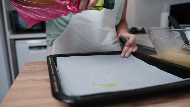 The pastry chef pours oil from a bottle onto parchment paper on a baking sheet, in slow motion.