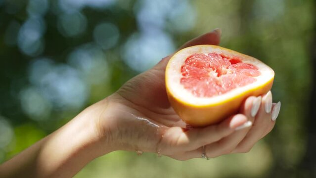 Piece of juicy grapefruit is squeezed out by graceful female hand with white manicure in garden in sunlight. Citrus juice flows down palm of hand, on hand is shadow from branches of trees.