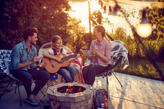 Little girl playing guitar by the fireplace, mom and dad clapping and singing along. Beautiful scene of family enjoying sunny summer day outdoors in the yard.