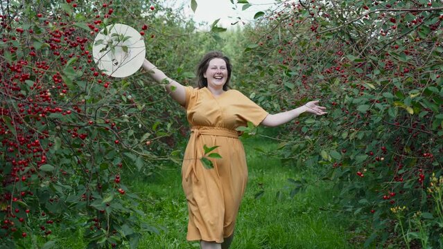 Plus size imperfect positive woman in bright dress runs through cherry orchard. Lady raises hat above her, smiles, enjoys the moment. Concept of self acceptance, mental health, imperfection of body.