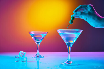 Portrait with glasses of classic dry martini cocktail with olives over colourful gradient background in neon glow. Bartender preparing cocktails