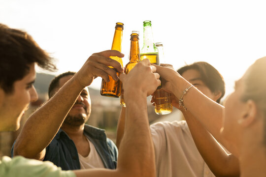 A group of unrecognizable young people toast downtown with glass bottles on the roof of an open-air apartment. A close-up of a multiracial group toasting with beers. Concept of multiracial youth.