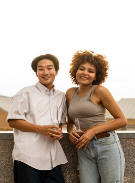 Vertical photo of a pair of multiracial friends are drinking beer on the rooftop of an outdoor apartment. The Korean boy and the girl with the afro hair look at the camera happily. Multiethnic friends