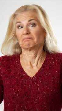 Vertical video portrait of happy senior mature woman attractive with blond hair wearing red sweater shrugging shoulders and gesturing I do not know isolated on solid white background.