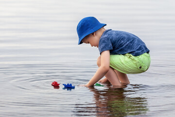 Kid boy playing with toy boat in sea water. Happy holiday by the sea. Happy kid playing with colour toy boats. Summer vacation. Cute little kid play and floats his toy ships.