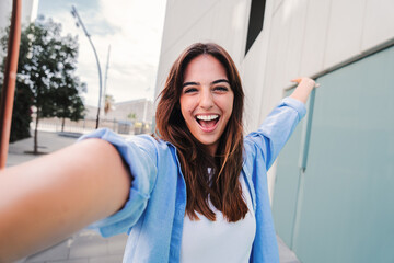 Fototapeta Happy young caucasian student lady looking at camera and taking a selfie portrait having fun, standing outside. Front view of laughing woman shooting a photo for social media at the university campus obraz