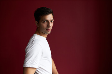 Profile view of fit sport coach in white t-shirt going to training session or gym posing against red studio background with copy space on the left, looking at camera with confidence