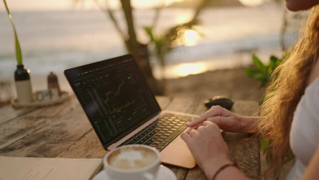 Female cryptocurrency trader at laptop checks charts online working remotely at outdoor tropical seaside cafe at sunset. Woman crypto broker analyses graphic of stock exchange rates by ocean close-up