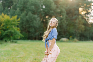 Young Caucasian woman or girl in a summer dress and a denim shirt is walking on the grass, holding...