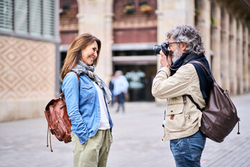 Portrait of casual mature woman posing smiling for photo taken with camera by husband on city street. Couple of happy tourists enjoying their retirement vacation in Europe. Love and romantic getaways.