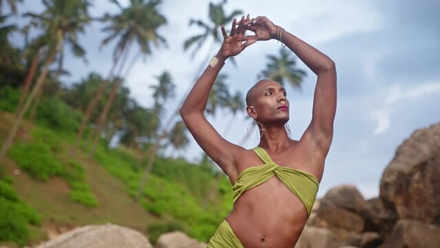 Gay bipoc man in open dress, boutique brass jewelry, makeup poses on a stone inside picturesque palm plantation. Homosexual ethnic elegant person in open outfit stands in jungle among rocks.