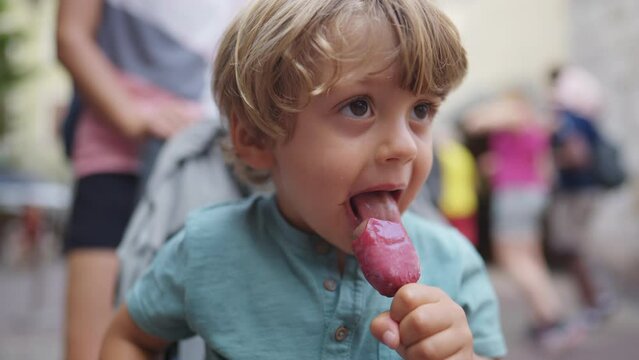 One cute small boy eating ice cream on stick portrait face close up. Kid snacking during summer day outside seated on stroller