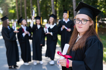 Fototapeta Portrait of a young caucasian woman in glasses and a graduate gown against the background of classmates. A group of graduate students outdoors. obraz
