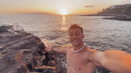 One attractive and happy young man at the beach smiling and looking at the camera holding and taking a selfie with his phone. Summer time in vacation holiday at the sunset
