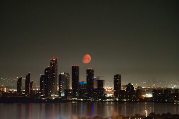 An early morning view of the moon setting behind buildings. The moon is a deep red because of smoke from wildfires in western Canada