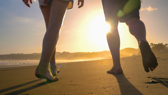 LOW ANGLE VIEW, CLOSE UP: Couple walking barefoot on sandy beach in golden light