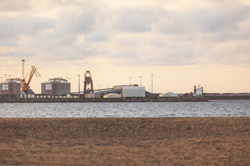Cranes in a port with energy resources, like hydrogen tanks and carbon, coal, and oil reservoirs for the energy and heavy industry, photo with orange sand and blue water of the ocean