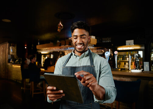 Happy young biracial man in apron pointing at digital tablet while working as waiter in bar