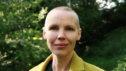 Bald middle-aged woman in a green coat on a background of green nature. Freedom, victory, chemotherapy concept.