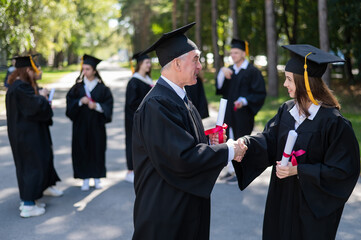 A group of graduates in robes outdoors. An elderly man and a young woman congratulate each other on their graduation.