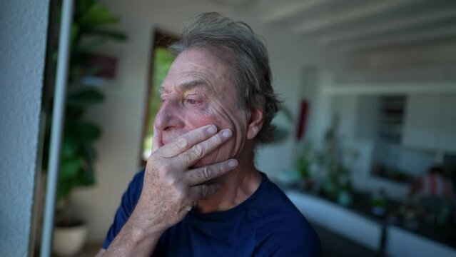 Concerned senior man with preoccupied expression. Close up face of an older male person with worried emotion standing by window at home with thoughtful look