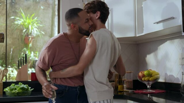 casal gay inter racial brasileiro na cozinha se beijando e abra&ccedil;ando com ta&ccedil;as de champanhe nas m&atilde;os no dia dos namorados