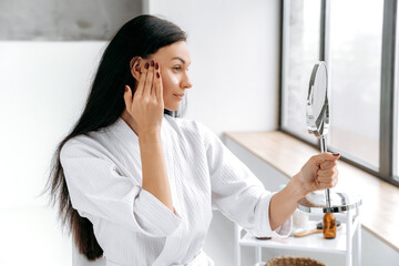 Using face cream. Happy caucasian woman, in bathrobe, holds a mirror in her hand, applies a...