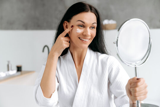 Face cream. Lovely well groomed caucasian woman, in bathrobe, holds a mirror in her hand, applies a moisturizing, anti-aging cream on her face, takes care of her skin and youth, smile