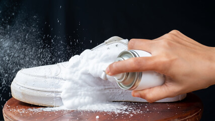 A woman sprays cleansing foam onto white leather shoes. Care for leather footwear.