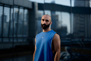 A man in a blue sleeveless shirt poses in warm, golden sunlight that accentuates his toned physique and thick beard. In the background, a reflective glass wall shows an outdoor fitness area.