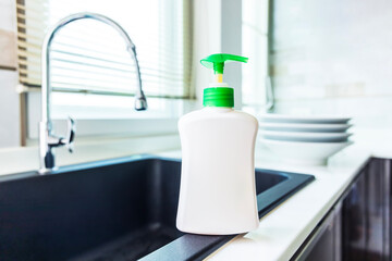 Dishwashing liquid bottle on kitchen sink and clean plates background.Dish cleaning liquid bottle with blank or white empty label space for text near window in modern design kitchen and water tap.