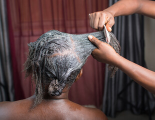 Hands of an African Nigerian stylist combing and applying relaxer cream to the  hair of a woman in...