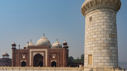 Obraz premium The white marble minaret of the Taj Mahal mausoleum against the blue sky. In the distance, the red sandstone Kau Ban mosque with domes and arches is visible. Blue sky. India. Agra