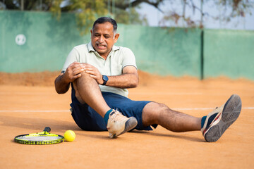 Indian fallen injured senior man suffering from knee pain while playing tennis at court - concept...