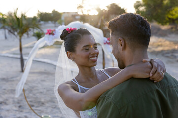 Caucasian newlywed bride embracing and looking at groom at wedding ceremony at beach