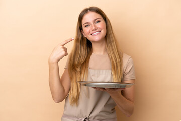 Young blonde waitress woman with tray isolated on beige background giving a thumbs up gesture