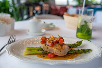 Close-up view of fried tuna steak with grilled asparagus, tomatoes and salt oyster sauce on white plate in seafood restaurant. Soft focus. Gourmet theme.