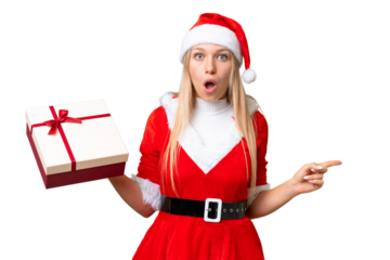 Young blonde woman with christmas hat and holding a gift over isolated chroma key background surprised and pointing side