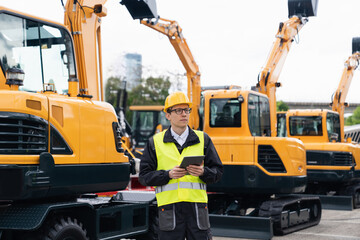 Engineer in a helmet with a digital tablet stands next to construction excavators	