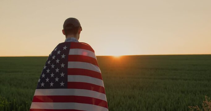 Young male farmer with usa flag on his shoulders looks at sunrise over wheat field