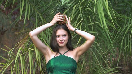 young woman holding a pineapple on her head against a jungle background