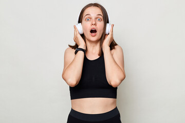 Shocked young Caucasian woman in black top listening music with headphones, looking at camera isolated on white background looking at camera with open mouth and big eyes.