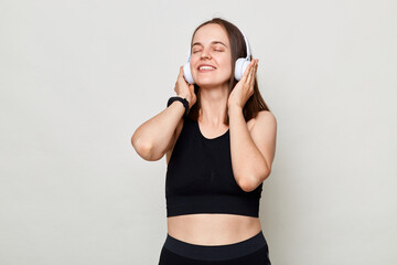 Photo portrait of pretty young girl touch earphones listen music toothy smile wearing black top isolated on white background enjoying favorite song.