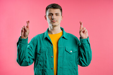 Young man praying with crossed fingers on pink background. Guy begs someone