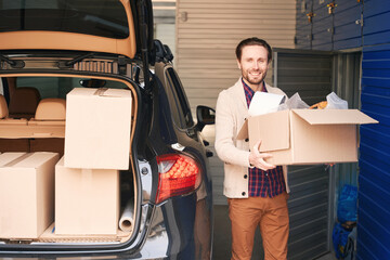 Smiling guy with cardboard box near the trunk of the car in a warehouse