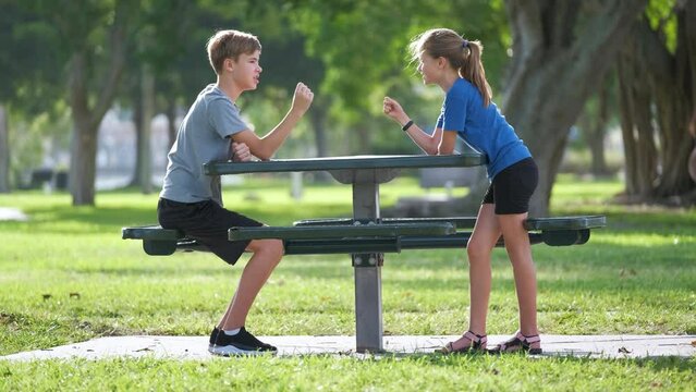 Two happy children boy and girl playing rock-paper-scissors sitting on outdoor bench in summer park on sunny day. Concept of competitive relations between children