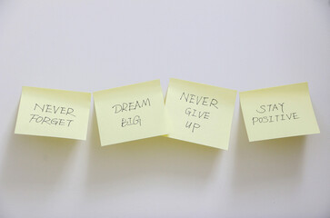 Four yellow sticky notes stuck across white wall with motivational and inspirational messages as a reminder to never forget to dream big, never give up and stay positive