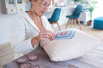 Portrait of happy serene woman working embroidery on a white pillow in living room at home sitting at the table and smiling. Renewal house decoration. Indoor leisure activity hobby at home. Art craft - obrazy, fototapety, plakaty